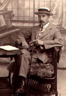 Sepia-toned Edwardian studio photograph, around 1915, portrait orientation. A middle-aged white man with a neat upturned moustache sits three-quarter facing the camera, wearing a flat-brimmed straw boater hat, a light tweed suit with waistcoat, and a wing collar. He rests both gloved hands on the crook of an upright walking cane held in front of him, sitting in a woven wicker chair with a fur or animal-skin draped over one arm. Behind him is a painted studio backdrop suggesting a leaded lattice window and drapery, with a small side table bearing a book at left. Soft, formal, dignified mood in warm brown monochrome tones.